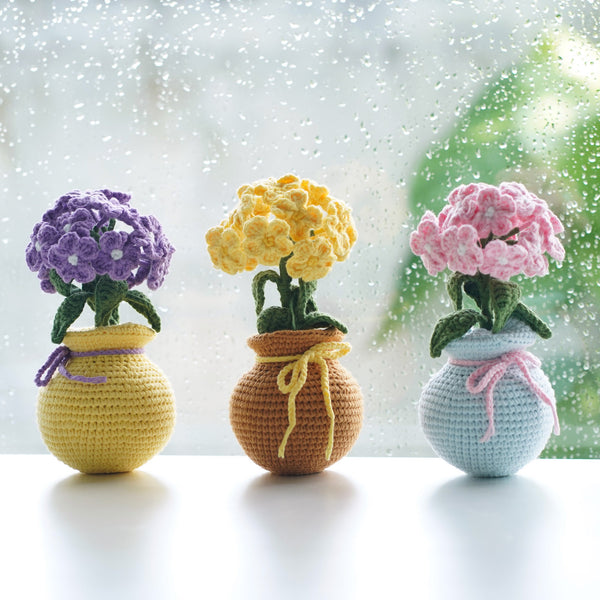 Three crocheted flower vases with flowers in front of a window with raindrops.