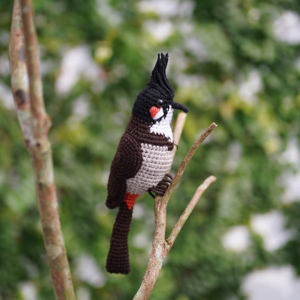 A crocheted red-whiskered bulbul bird perched on a branch with green leaves in the background.