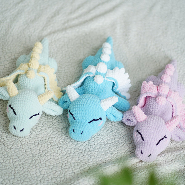 Three crochet plush toys shaped like baby dragons in blue, beige, and pink colors, made from chenille yarn, displayed on a soft surface.