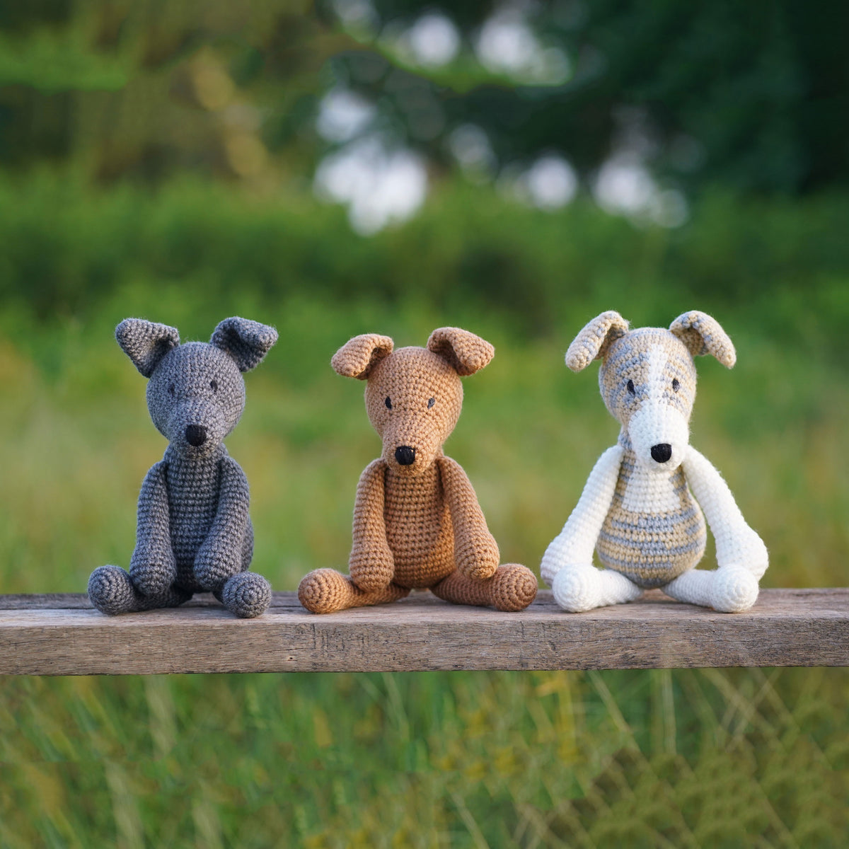 Three seated crochet greyhound dogs in gray, tan, and pale striped colors on a wooden rail