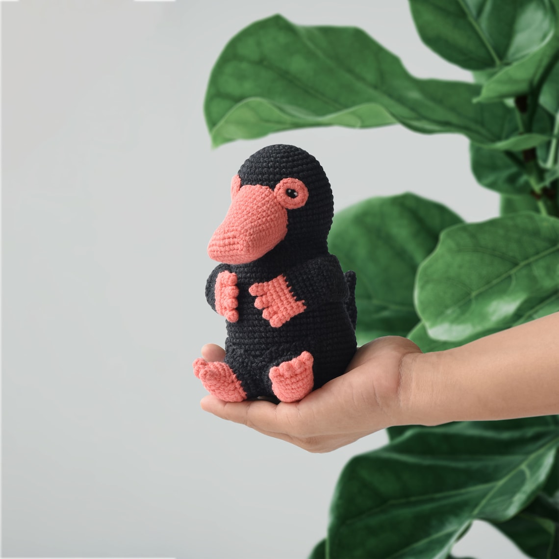 Black crochet platypus with a coral-colored bill and feet held in a hand beside green leaves