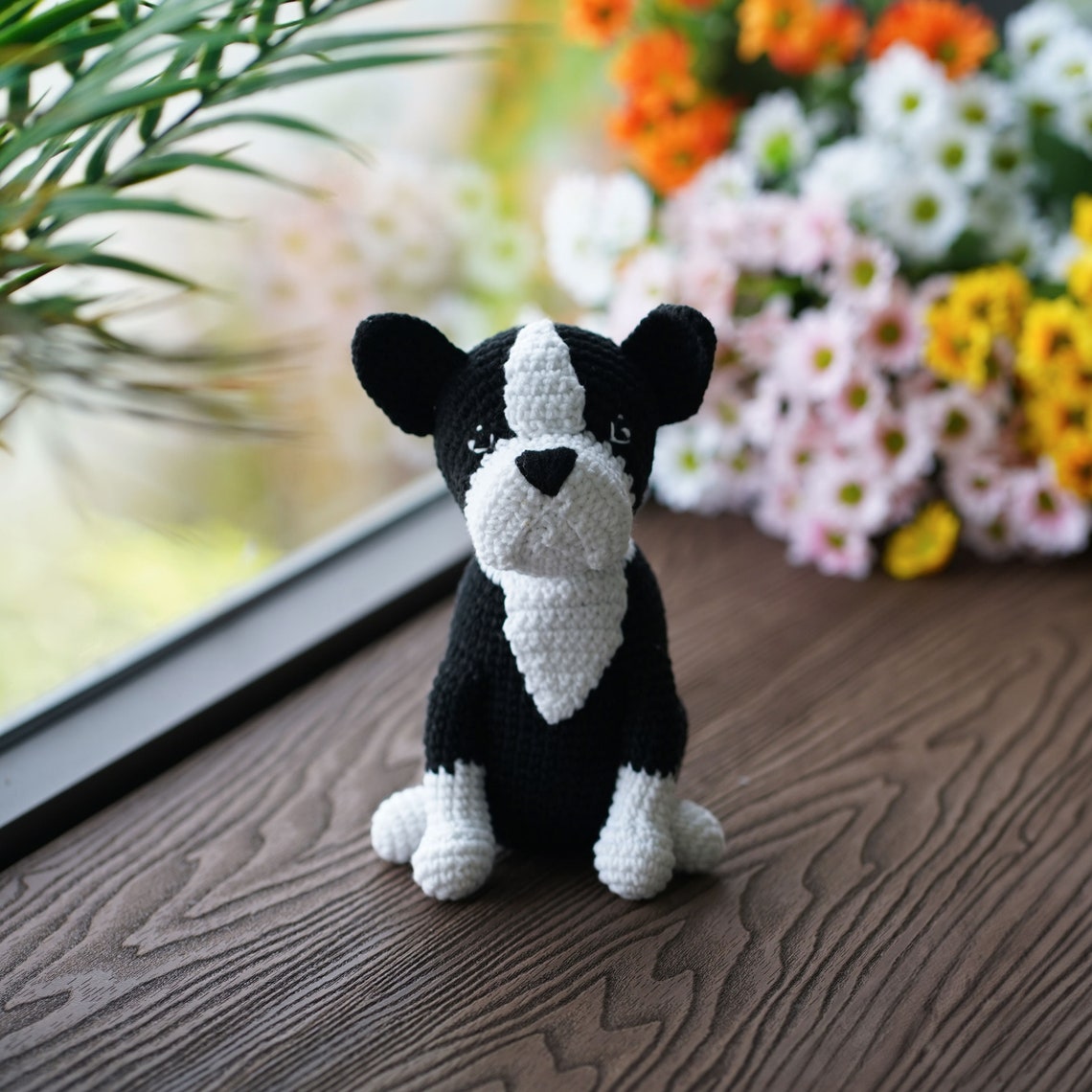 Black-and-white crochet Boston terrier puppy sitting on a wooden table beside flowers