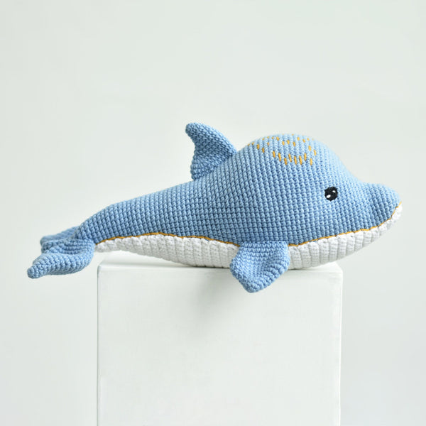 A crocheted plush dolphin toy in light blue and white, resting on a white pedestal against a clean white background. The dolphin has a small embroidered yellow smiley face on its forehead and a black bead eye.