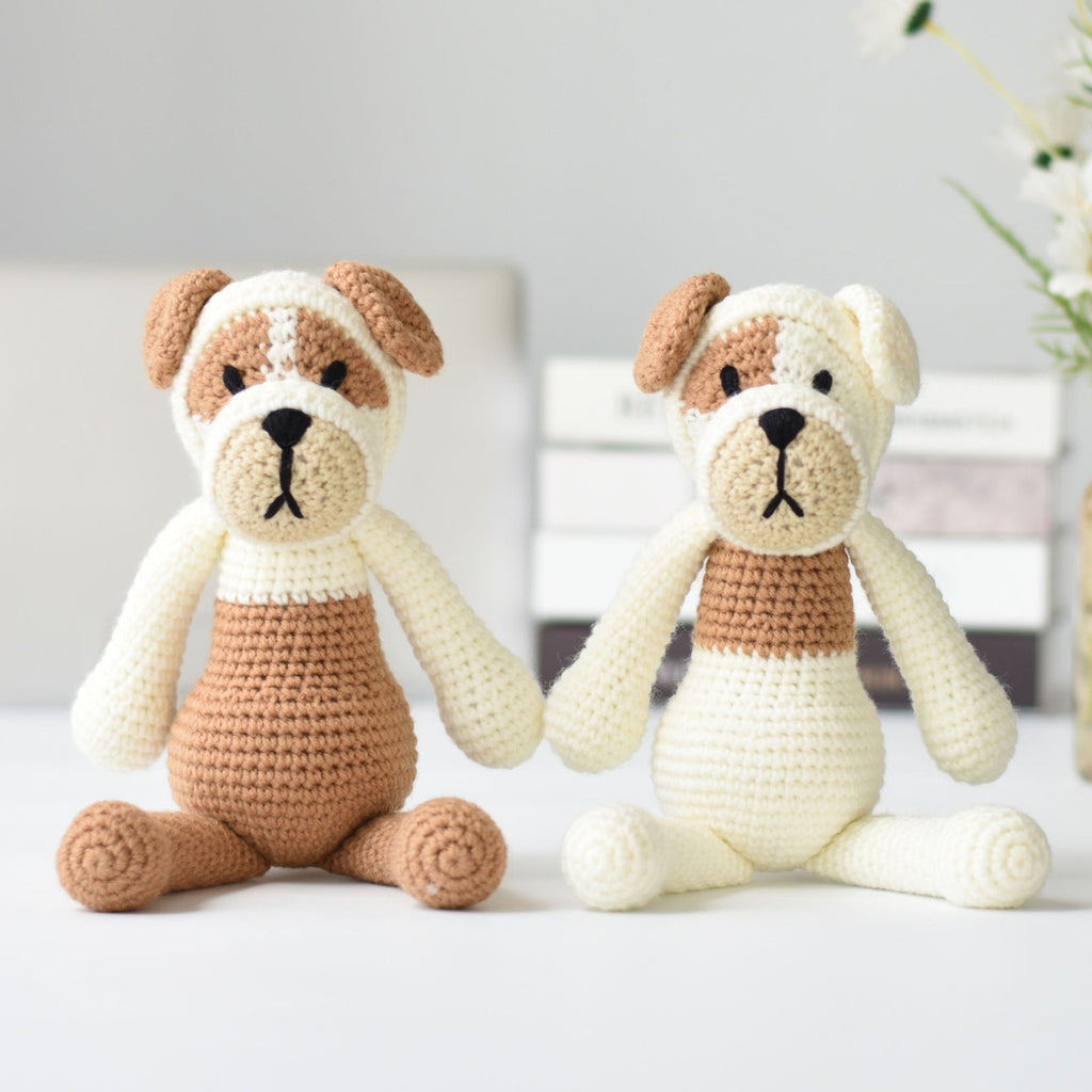 Two crocheted amigurumi dog toys in cream and tan are displayed on a white surface with a soft-focus background containing books and a vase of white flowers.