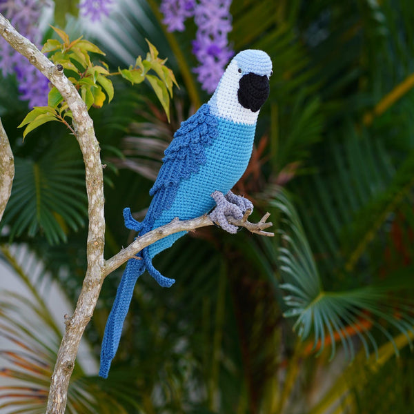 A crocheted blue parrot with a black beak and gray feet perched on a tree branch, set against a lush background of green palm leaves and purple wisteria flowers.