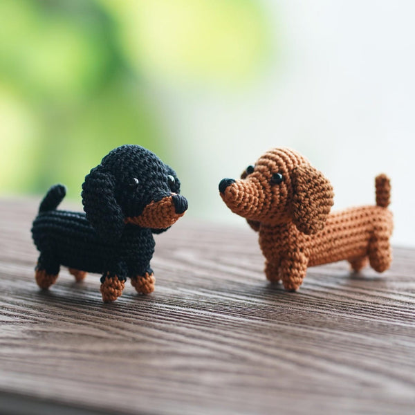 Two small crocheted dachshund toys, one black with tan markings and one tan with black markings, standing on a wooden surface with a blurred green and white background.