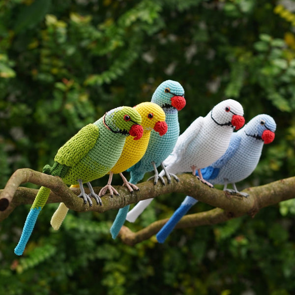 Four crocheted parrot birds in green, yellow, light blue, white, and dark blue, perched together on a tree branch with a blurred green foliage background.