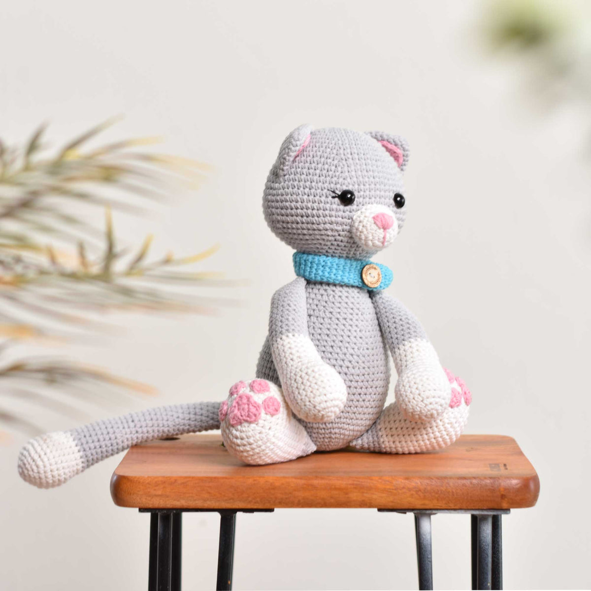 Seated gray crochet cat with a turquoise collar, pink nose, pink paw pads, and a long tail